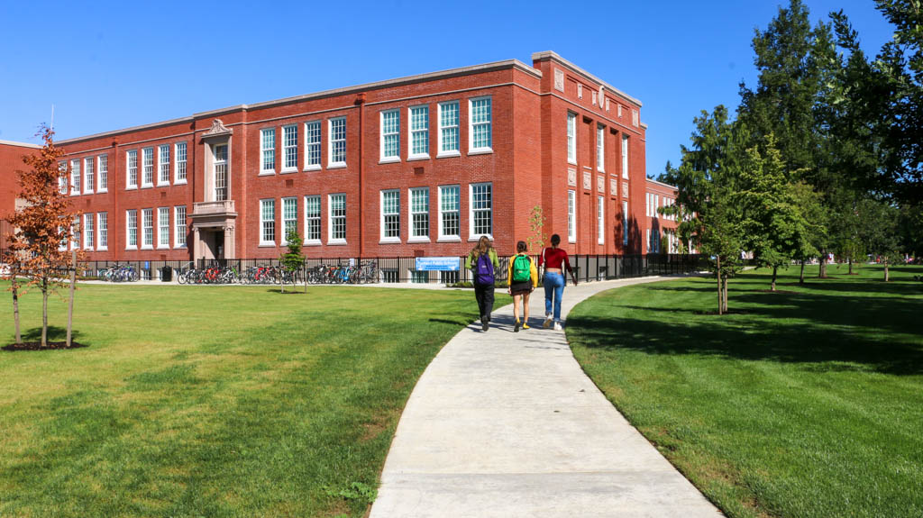 A three-story red brick school building with a green courtyard divided by a light gray sidewalk. Three students are walking away from the camera towards the school.
