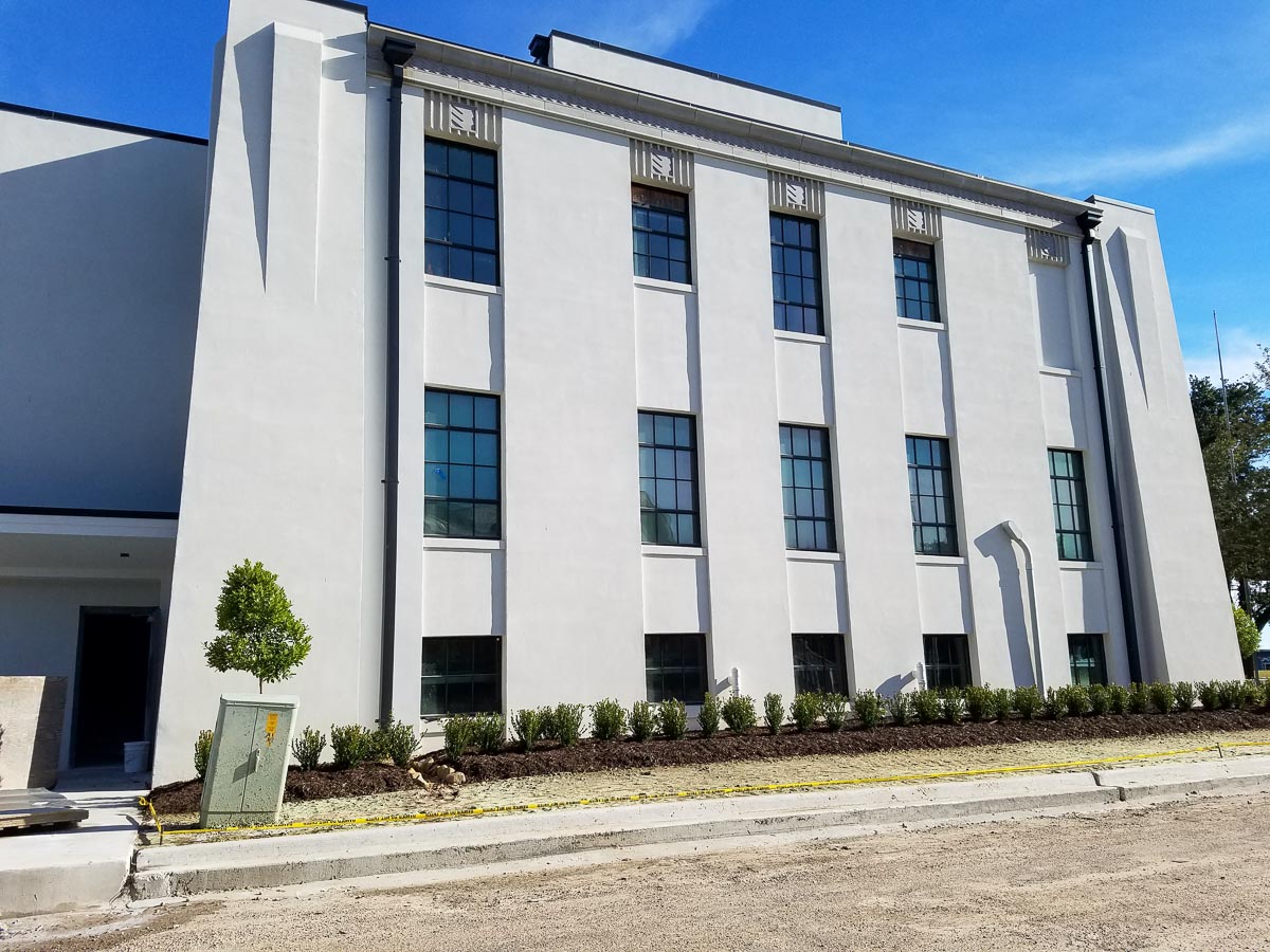 This large commercial building is painted white. There are shrubs lining the side of the building. 