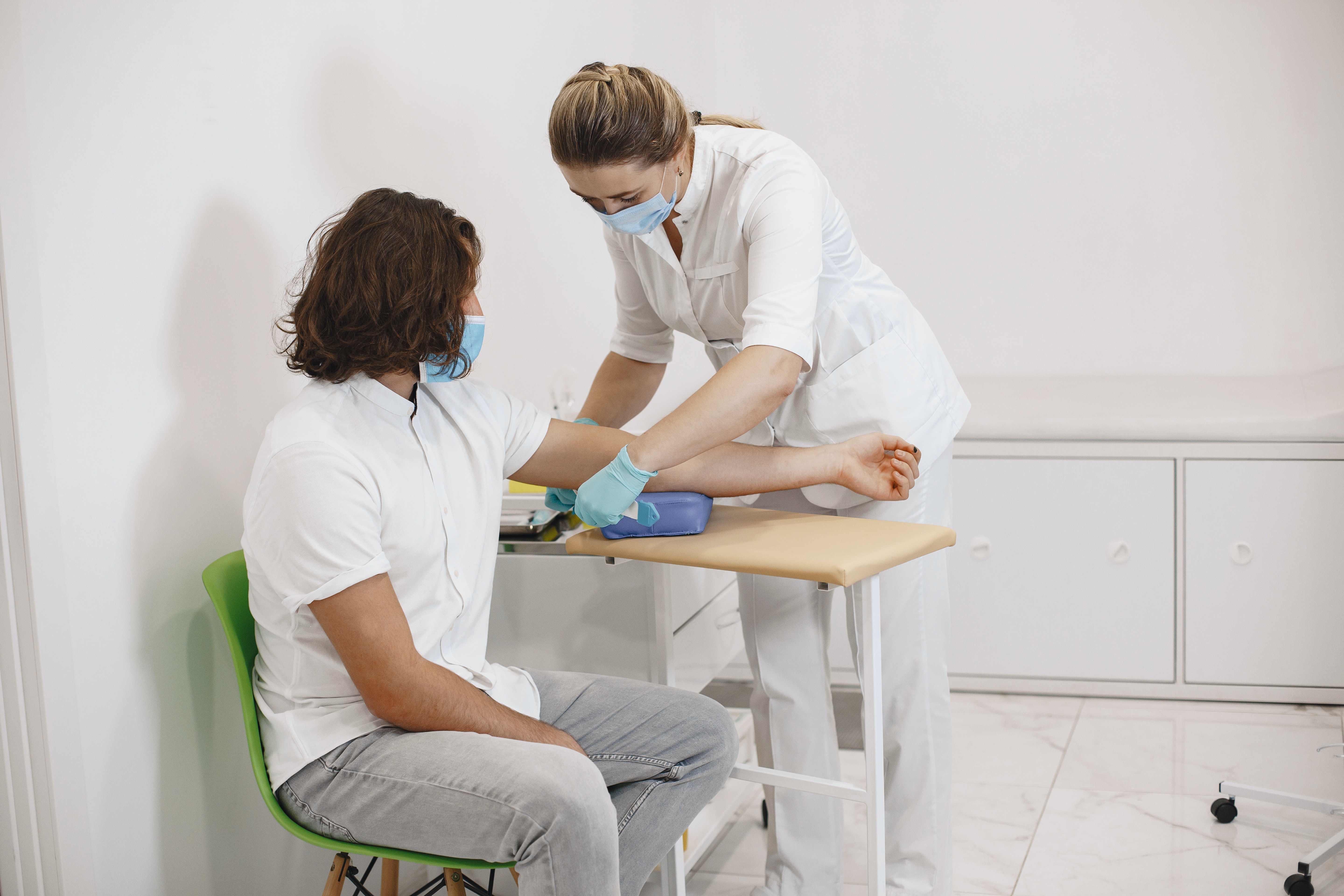 A nurse preparing a seated patient to draw blood. Both are wearing blue masks and white shirts.