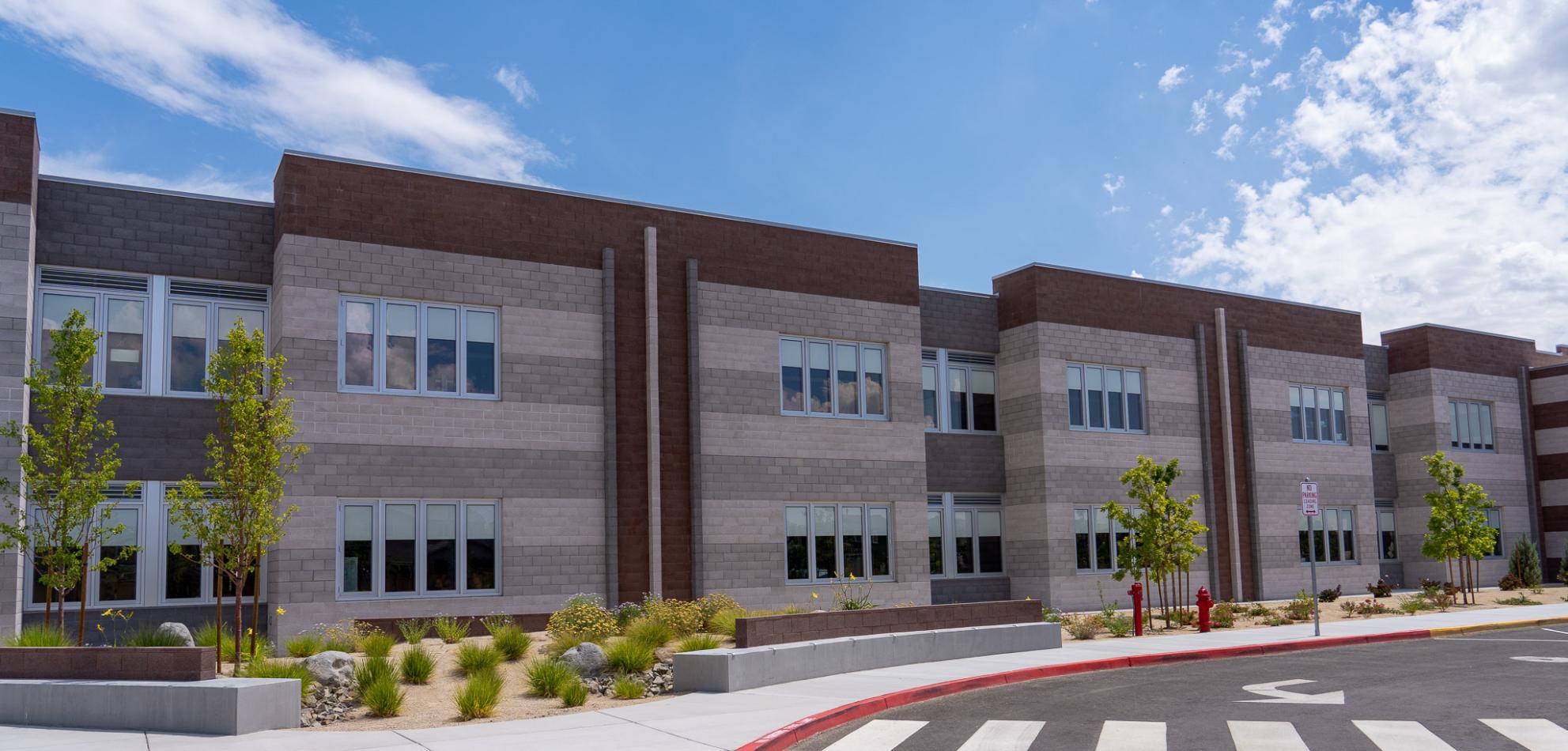 Students in the Washoe County School district in Reno, Nevada benefit from the abundant natural light conditions in multiple ways. The automated Transira shading system controls light and significantly improves occupant comfort. A two-story school building with brick in shades of gray and tan. The sidewalk in front is landscaped with small green bushes and trees.