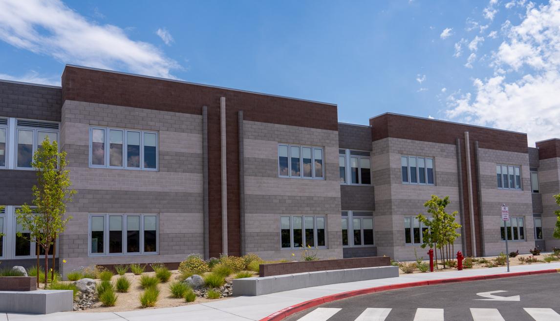 A two-story school building with brick in shades of gray and tan. The sidewalk in front is landscaped with small green bushes and trees.