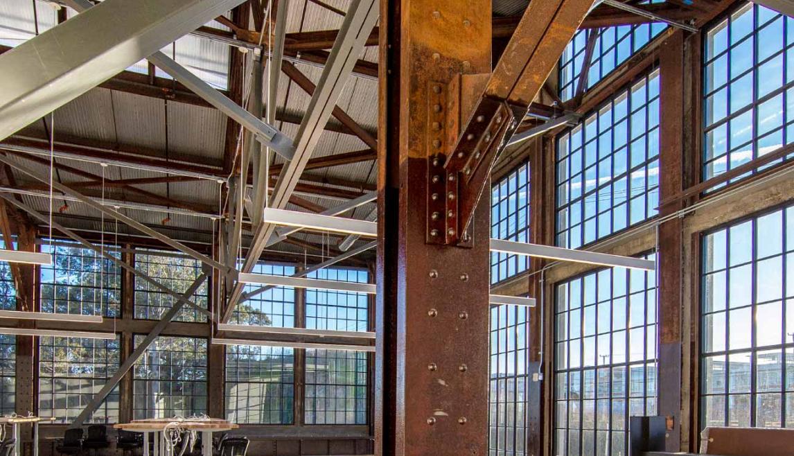 WINCO provided more than 48,000 square feet of its Series 3250 Steel Replica windows fitted with ¼” monolithic tempered clear glass. In addition to stunning aesthetics, the windows can withstand 110 mph winds as well as seismic activity. A large indoor room featuring a prominent metal beam, with natural daylight illuminating the space. The floor is visible beneath the beam, showcasing materials like steel and aluminum in the structure.