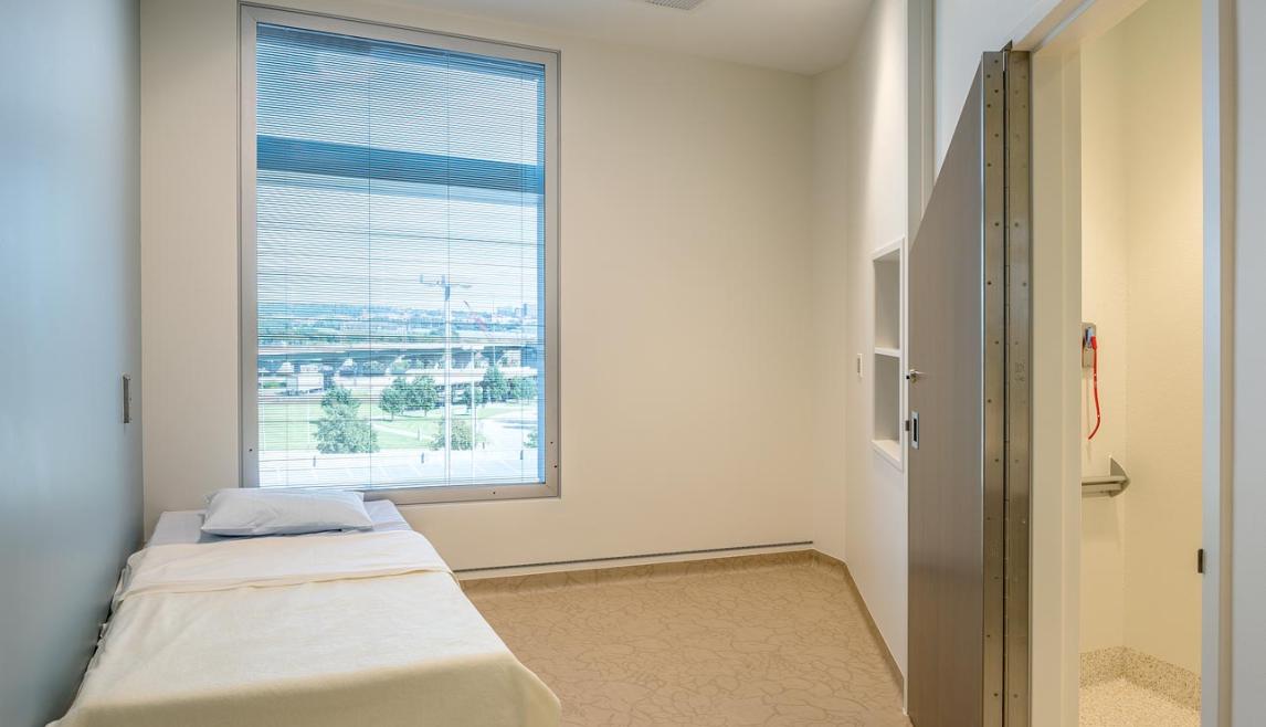 A behavioral care patient room with a bed and a window, equipped with human impact resistant windows for safety. 
