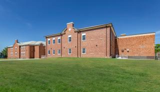 Exterior brick building featuring windows with a grassy field in front and blue sky in the background.