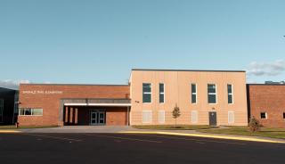 Modern school building with beige and brick facade under a clear blue sky. Manicured lawn and young trees create a peaceful, sunny atmosphere.